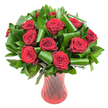 A luxurious, rounded bouquet of a dozen or more deep crimson roses sits like a jewel in a tall, translucent ruby-red glass vase. Each rose is shown in full bloom, the velvety petals tightly spiraled at the centre and unfolding outward in layers of intense, wine-dark red that catch the soft, even studio light. The blooms are densely clustered to form a generous dome, their rich colour set off by an abundance of dark green foliage. Glossy, flat leaves-reminiscent of Ruscus or Salal-create a dense, verdant backdrop while broader, emerald Aspidistra-style leaves have been expertly rolled and curled into sculptural loops and cylinders. Those folded greens frame and cradle the rose heads, adding a modern architectural rhythm and generous volume to the composition. The vase is slender and elegantly shaped, with a faint ribbed or twisted texture that throws delicate vertical highlights and echoes the roses' deep hue; its translucent surface softens the stems' silhouettes and anchors the arrangement with quiet sophistication. Set against a crisp white background, the colours read with clear contrast and clarity: the red feels almost tactile and the greens gleam. Standing as a romantic centrepiece or an anniversary gift, the image conveys classic passion tempered with contemporary design. Imagine the faint, timeless scent of newly opened roses and the comforting weight of the vase in hand-an arrangement that would suit a candlelit table in a Highgate flat near Waterlow Park or a polished dining room by Highgate Hill, where warmth and thoughtful craft meet.
