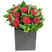 A contemporary bouquet built around rich, velvety crimson roses that catch the eye with their generous spirals of petal-each bloom densely layered and almost sculptural against a backdrop of glossy, artfully curled green foliage. The leaves are looped and folded with purposeful rhythm, creating overlapping waves of emerald that both support and set off the roses' deep red tones. The composition sits in a low, matte-black square vase whose solid geometry grounds the arrangement and gives the crimson blooms a bold architectural stage. Bright, even studio-style lighting in the photograph picks out highlights on each leaf and the subtle sheen of the rose petals, suggesting the freshness of a just-opened bouquet and the clean, modern feeling of a gift prepared for a city setting. As a florist who often brings flowers to Highgate Village and to tables near Highgate Pond, I picture this piece on a sleek sideboard in a Hampstead Heath-facing flat or as a romantic centerpiece for an intimate anniversary dinner on Swains Lane. Close to the blooms you can almost imagine the scent-a classic rose perfume, warm and heady, lifted by the green, almost tropical notes of the foliage. This arrangement is intentionally poised for moments that call for affection and quiet drama: a declaration of love, a thoughtful thank-you, or a way to turn an ordinary room in Highgate into something quietly unforgettable. The craftsmanship reads as confidence: restrained, elegant, and utterly expressive in crimson and green.
