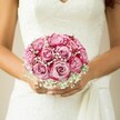 This bridal bouquet, nestled against a lace-adorned wedding dress, tells a story of Highgate romance and celebration. Vibrant roses, their petals spiraling from blush pink hearts to radiant fuchsia outer tips, create a crescendo of colour that dazzles in the soft daylight filtering through local chapel windows. Wisps of baby's breath flutter among the roses, offering a feathery white glow that softens the arrangement and imparts the gentle innocence of a perfect English morning. The bouquet is cradled in warm, careful hands, each finger accented by a touch of pale, glossy polish-the small, human details that matter most on a wedding day. A graceful sash ripples just above where the bride's palms meet, introducing texture and movement. Every bloom, every fold of lace and gleam of petal, seems imbued with the spirit of a Highgate celebration-timeless, joyful, and deeply personal. This arrangement is not just a bouquet; it's a memory waiting to blossom, thoughtfully designed by a florist who understands the nuance and charm of North London weddings.