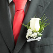 In cool, even light the ivory rose sits proudly in the lapel, its velvety petals opening with a calm dignity that draws the eye against a smart charcoal jacket. This particular composition pairs the bloom with tiny clusters of baby's breath, creating a frothy counterpoint that softens the rose's form and suggests purity and gentle devotion. Beneath, fine slivers of fern and curled greenery arch elegantly, introducing movement and depth while their darker tones frame the central flower. The boutonnière is pinned over the checked texture of a shirt and a bold red silk tie, a classic contrast that balances formal flair with restrained floral romance. A narrow white ribbon is tied at the base, its neat bow catching a whisper of light and completing the piece with a ceremonial whisper rather than a shout. The scent is imagined - a cool, dewy rose note mingled with herbaceous green - like lingering floral freshness after a morning run through Highgate's lanes or a stop at the local flower stall off Highgate High Street. As a local florist, I assemble this with attention to proportion and wearability: wiring the stem for secure pinning, trimming the foliage so it sits flat, and flexing the bow to move with the jacket. Flowers Highgate often creates pieces like this for ceremonies in nearby venues and for couples who appreciate subtlety, craftsmanship, and a touch of village charm on their special day.