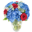 A luminous bouquet sits in a clear glass vase against a clean white backdrop: a dense cloud of sky-blue hydrangeas forms the soft, voluminous heart of the arrangement, each tiny floret overlapping to create a plush, almost pillow-like texture. Woven through that cool blue mass are three velvety red roses, their petals still tightly cupped and rich with a deep crimson that reads as both intimate and celebratory. Sprigs of delicate white baby's breath drift between the larger blooms like soft, ivory threads, adding an airy, starry contrast that lightens the composition. Rounded eucalyptus leaves in muted sage-green nestle through the blooms, their matte surface and subtle herbal scent implied in the arrangement, giving structure and a gentle, restorative aroma in the mind's eye. The vase itself has a softly rounded base and a slightly tapered neck; its transparency reveals the healthy green stems and the clarity of the water, a visual promise of freshness. Bright, even lighting highlights the saturated blues and reds and casts gentle, minimal shadows that keep the focus entirely on texture and colour.As a florist familiar with Highgate and the nearby stretch toward Hampstead Heath, I imagine this bouquet placed on a sunlit kitchen table in Highgate Village or at a small celebration in Pond Square-an arrangement that reads as both contemporary and reassuringly classic. The combination of cobalt hydrangea and romantic red rose makes a bold but tasteful statement, suitable for anniversaries, table centrepieces, or to convey affection with an elegant, local sensibility. The overall effect is cool and uplifting, a confident display that soothes with its soft forms and stirs with its passionate red notes.