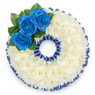 A bespoke sympathy wreath formed into the distinct shape of the numeral '0', photographed from above against a clean white backdrop. The arrangement is densely packed with hundreds of small white spray chrysanthemums, their tiny petals overlapping into a soft, pillowy surface that reads visually as pure and consoling. A narrow pleated ribbon in deep royal blue traces both the outer and inner edges of the zero, its satin sheen catching a cool, diffused light to create a scalloped, formal frame. On the upper left curve, a trio of vivid royal blue roses-each with petals unfolding from tight centres into layered spirals-creates a focused, emotive accent. Glossy dark green leaves are tucked behind the roses, lending contrast and a subtle scent of fresh foliage against the neutral chrysanthemum base. The overall composition balances quiet brightness with a respectful, dignified silhouette, a design often chosen to mark an age or special number at a memorial service. The blue ribbon and roses may signify a connection to a uniform, a favourite colour, or a particular association; in Highgate we sometimes craft number tributes like this for families remembering a loved one whose life was closely tied to a club, regiment, or cherished pastime. Soft studio lighting produces gentle shadows between the blossoms, giving the circular form gentle depth while keeping the focus on texture and tone. As with all funeral pieces, the tactile detail-the ruffled ribbon, the cushiony chrysanthemums, the velvet of the blue rose petals-intends to offer comfort, a quiet visual embrace for those gathered to remember.