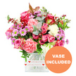 The photograph captures a lush, romantic bouquet arranged in a clear, square glass vase, with every detail standing out crisply against a spotless white background. The arrangement is a tapestry of pinks and purples: large roses in deep fuchsia, magenta, and soft blush form the core, their petals layered thickly and some edged with deeper tones that give them a luxurious, almost velour appearance. Nestled between the roses are generous carnations in coral-pink, salmon, and light peach, their ruffled petals tumbling outward and contributing a soft, cloud-like fullness. Punctuating the bouquet are small daisy-style chrysanthemums in lavender and lilac shades, each blossom finished with a bright green centre that lights up the display and adds a contemporary freshness. A scattering of creamy white and paler blooms keeps the arrangement airy and balanced, preventing the pinks from feeling too dense. The greenery is abundant yet refined, with rounded silvery eucalyptus leaves, darker glossy foliage, and a few reddish-brown accents echoing the seasonal tones often seen around Highgate's leafy avenues. Wrapping the base of the vase is a neat white band, printed repeatedly with little red hearts and the tender message all my love, clearly marking this as a gift intended from the heart. In the bottom right corner, an orange circular emblem reads VASE INCLUDED in clear white capital letters, indicating the bouquet will arrive already styled and ready to stand on a table. Created with the care you would expect from Flowers Highgate, the overall effect is warm, inviting, and ideal for same-day delivery to homes near Highgate Village, Waterlow Park, or the surrounding North London neighbourhoods.