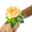 A romantic wrist corsage rests gently on the inner forearm, captured against a clean white background so every texture and hue reads clearly. At the heart of the design sits a single, fully opened rose in a soft peach-to-apricot tone, its velvety petals spiraling inward with a warm, luminous glow that suggests a quiet sunrise. Nestled below and to the left, two bright lime-green button chrysanthemums - compact, spherical pom-poms - add a lively, tactile contrast; their dense, spiky florets feel almost like laughter captured in bloom. Rich, glossy foliage forms a deep, structured base: broad dark-green leaves, likely salal or ruscus, fan beneath the flowers and create depth against the skin. A long, variegated leaf with pale stripes is artfully folded and curled, sweeping to the right and lending an architectural, contemporary line that balances the softness of the rose. The corsage is pinned or banded snugly at the wrist, though the fastener is hidden; the composition reads as fresh, meticulously assembled, and ready for a prom, wedding or evening celebration. Light seems soft and diffuse, suggestive of a late-morning preparation in a Highgate flat near Waterlow Park, the faint perfume of rose mingling with green, cut foliage. As a local florist's piece, this wristlet expresses tender elegance - quiet, wearable, and crafted to accompany vows, first dances, or the small, bright moments of a day remembered. It combines refined colour harmony with a playful pop of lime, offering both tradition and a modern, architectural twist.