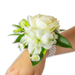 A delicate wrist corsage of pristine white blooms rests gently on a fair-skinned wrist, photographed against a clean white backdrop so every texture and subtle tone reads clearly. At its heart sits a creamy ivory rose, petals unfolding in a soft spiral that catches the light with a warm, velvety sheen; beneath and slightly to the left a trumpet-shaped white alstroemeria (Peruvian lily) opens its ribbed petals to reveal a pale green-yellow throat that gives the composition depth. Sprigs of airy baby's breath thread a misty softness through the arrangement, while tiny white daisy-like florets with bright golden centers add a cheerful, refined punctuation near the wrist. Several green buds-fresh and taut-promise future blooms, their vivid chartreuse tips echoing the alstroemeria's throat. Broad, glossy deep-green leaves cradle the flowers, their smooth, waxy surfaces forming a rich contrast to the delicate whites and lending the piece a just-cut-from-the-garden freshness. The flowers are assembled atop a chic multi-strand faux-pearl bracelet, each round pearl luminous and cool against the skin, the elastic strands looping comfortably around the wrist to secure the floral cluster. Imagine the faint, clean scent of the rose and a whisper of green leaf when the wrist moves-an understated fragrance that evokes early-morning walks through Highgate Village or along Waterlow Park. The overall effect is one of quiet elegance and handcrafted care, a petite floral statement suited to weddings, proms, and special evenings where simplicity and refinement are the lasting memory.