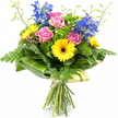 A colourful, contemporary bouquet is presented upright against a clear white background, its shape and colours crisply defined, as though waiting on a Highgate doorstep for the recipient to open the door. Several open pink roses form the gentle centrepiece, their soft, velvety petals spiralling outward and adding a note of tender romance. Around them, bright yellow gerbera daisies with deep brown centres stand like cheerful medallions, their daisy-like faces lending the design a sense of optimism and warmth. Stretching above the main cluster, elegant stems of vivid blue delphiniums provide vertical interest, each stem lined with small individual florets that create a graceful, almost architectural silhouette. The bouquet is generously framed by rich greenery: feathery fern fronds trail from the lower edges, softening the outline, while broad, glossy tropical-style leaves, some subtly striped, are artfully wrapped around the base of the blooms to form a smooth, structured collar that keeps the bouquet tidy and rounded. Threaded through the arrangement, clumps of bright, textured green filler foliage give the appearance of a lush, moss-like cushion, enhancing both volume and depth. A few slender, light green stems with tight buds gently emerge above the main flowers, hinting at blossoms still to open. Below the foliage, the straight green stems are neatly gathered and tied with a simple ribbon, creating the classic hand-tied look favoured by many Highgate clients. The overall impression is one of bright, joyful colour and careful craftsmanship, perfect for birthdays, thank-you gifts, or as a fresh floral accent for homes near Highgate Woods and surrounding leafy streets.